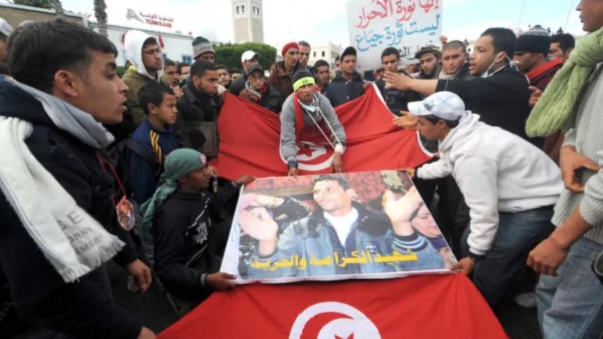 ‘আরব বসন্ত’ আন্দোলনের পথিকৃত মোহাম্মদ বোয়াজিজি’র পোস্টার ও তিউনিসিয়ার পতাকা হাতে ক্যাসেরিন অঞ্চলের মানুষ সরকারী রাজপ্রাসাদের সামনে বিক্ষোভ করছেন। ফাইল ছবি: এএফপি (২৮ জানুয়ারি, ২০১১)   
